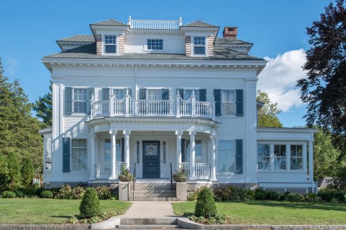 A large, elegant white house with a balcony, surrounded by greenery and a clear blue sky.