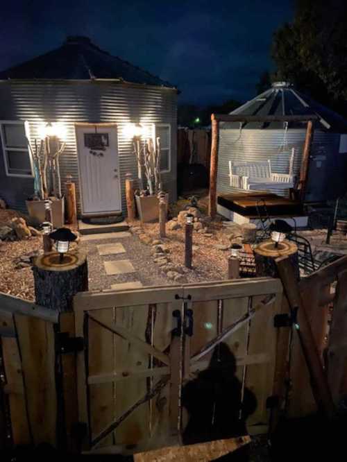 A cozy outdoor space at night featuring a round building, wooden gate, and seating area surrounded by lights and landscaping.