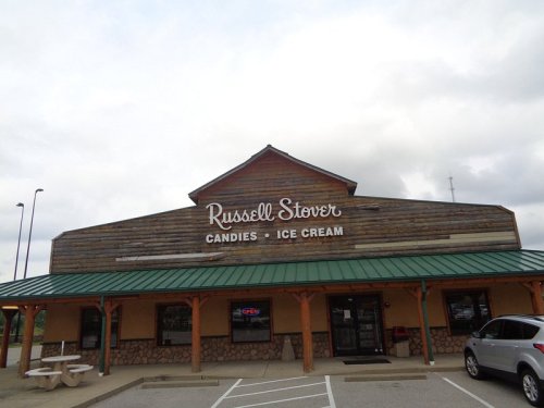 Exterior of a Russell Stover Candies and Ice Cream shop with a wooden facade and green roof under a cloudy sky.