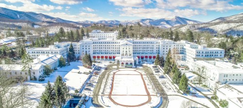 Aerial view of a large white building surrounded by snow-covered mountains and trees, with a red-roofed entrance.