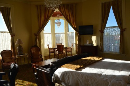 Bright hotel room with a large bed, elegant furniture, a dining area, and windows letting in natural light.