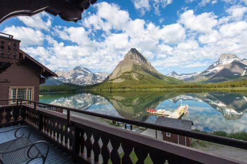 Scenic view from a balcony overlooking a calm lake, mountains, and a blue sky with fluffy clouds.