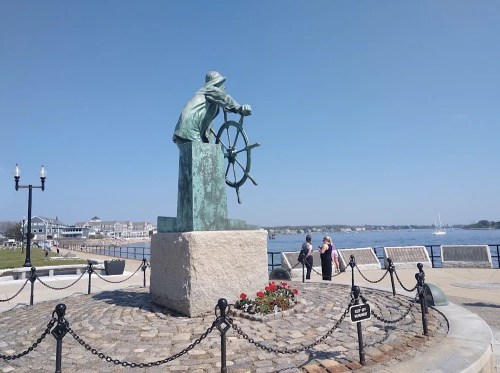 A bronze statue of a sailor at a ship's wheel, overlooking a waterfront with flowers and people in the background.