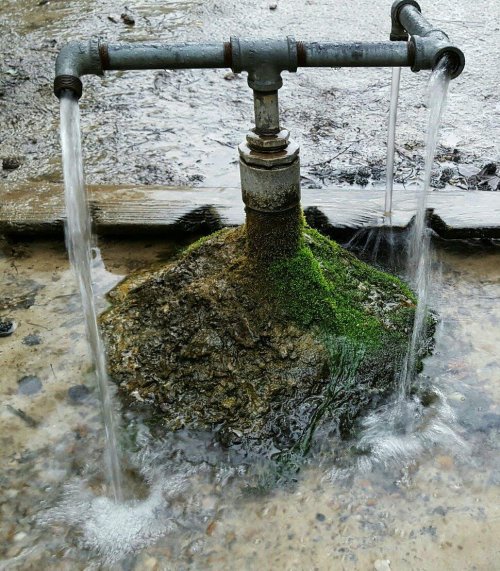 A metal pipe fountain with water flowing from two spouts, surrounded by moss and rocks.