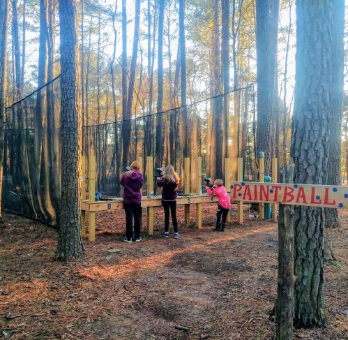 Three children prepare for paintball in a wooded area, with a sign indicating the paintball zone nearby.