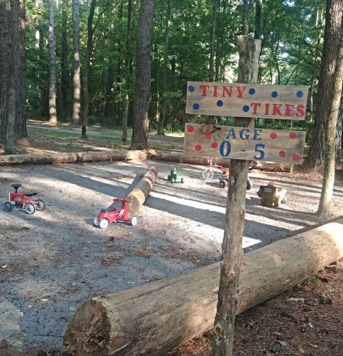 A playground area with toy vehicles for young children, marked by a sign reading "Tiny Tikes, Age 0-5" among trees.