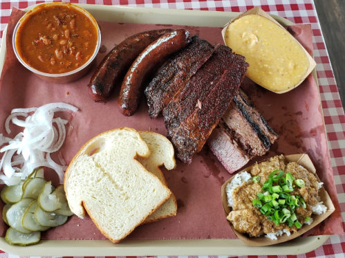A tray of barbecue featuring ribs, sausage, brisket, sides of beans, coleslaw, pickles, onions, and slices of bread.