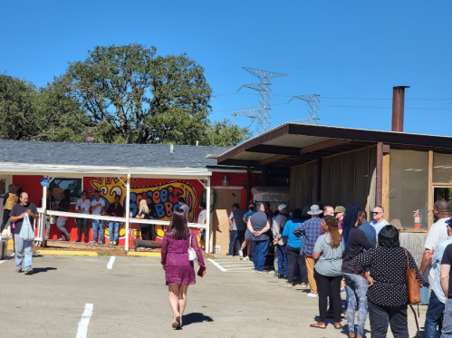 A long line of people waiting outside a colorful restaurant on a sunny day. Trees and power lines in the background.