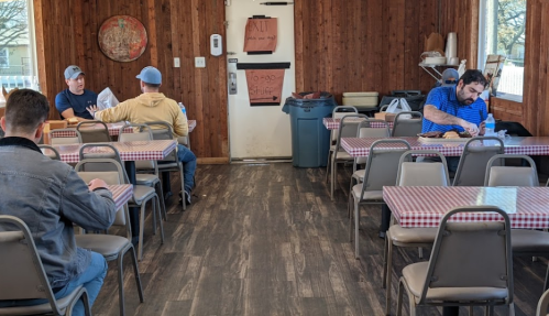 A casual dining area with wooden walls, checkered tablecloths, and two patrons seated at separate tables.