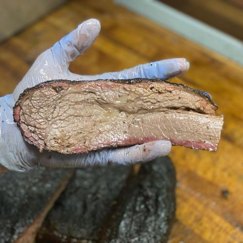 A gloved hand holds a thick slice of smoked brisket, showcasing its juicy texture and smoky crust.