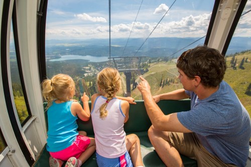 A man and two young girls ride a gondola, pointing at the scenic view of mountains and a lake below.