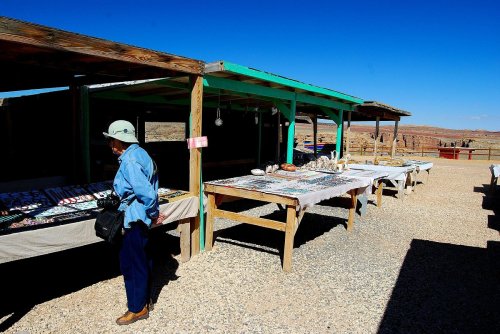 A woman browses tables of crafts and jewelry at an outdoor market under a clear blue sky.