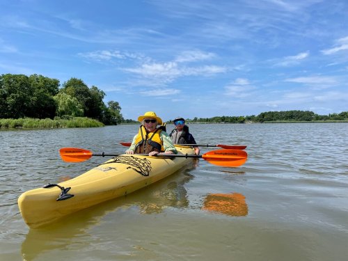 Two people in a yellow kayak with orange paddles, enjoying a sunny day on a calm river surrounded by greenery.
