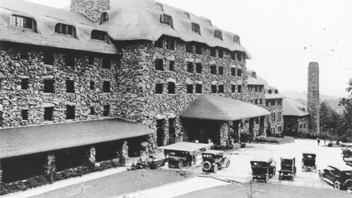 Historic stone hotel with a thatched roof, surrounded by vintage cars, set in a scenic landscape.