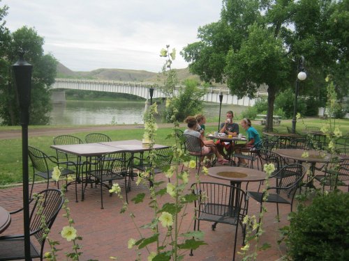 A group of people sitting at a table in a park by a river, with trees and a bridge in the background.