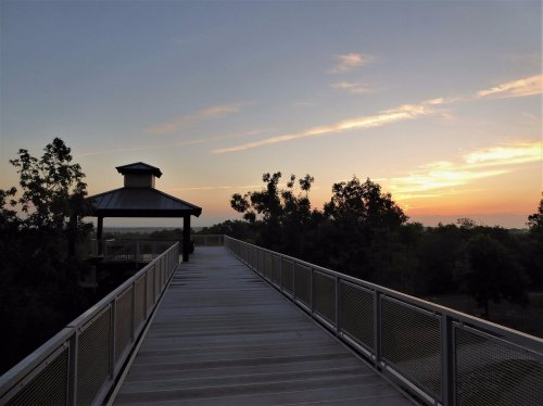 A wooden walkway leads to a gazebo at sunset, surrounded by trees and a colorful sky.