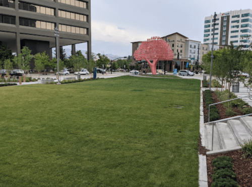 A green park area with a pink tree sculpture and modern buildings in the background under a cloudy sky.