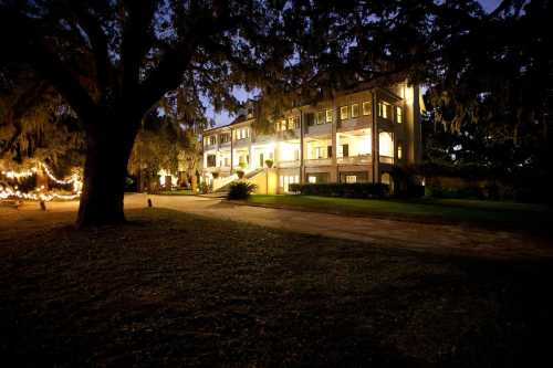 A large, illuminated house surrounded by trees at dusk, with a pathway leading up to it.