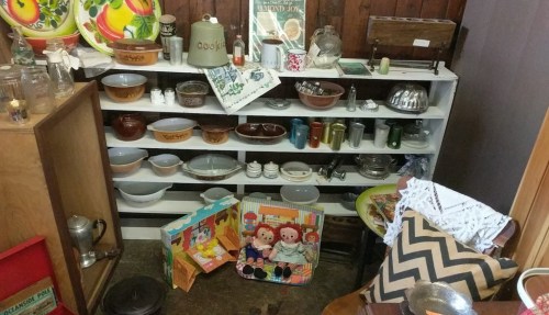 A vintage display of various kitchenware, bowls, and toys on a white shelf in a cozy shop setting.