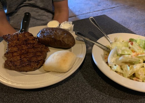 A plate with a grilled steak, baked potato, roll, and a side salad on a table.