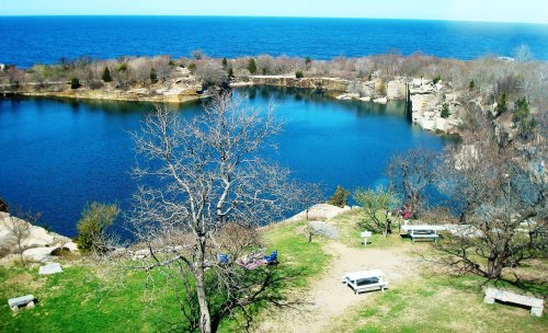 A serene view of a blue lake surrounded by rocky shores and bare trees, with the ocean in the background.