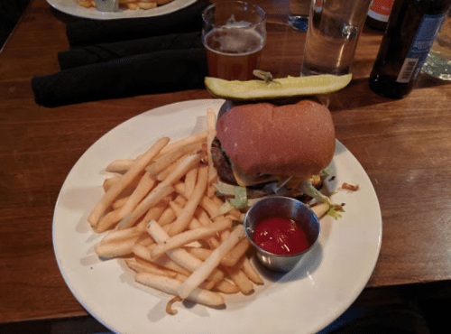 A plate with a cheeseburger topped with a pickle, served with fries and a small cup of ketchup, alongside a beer.