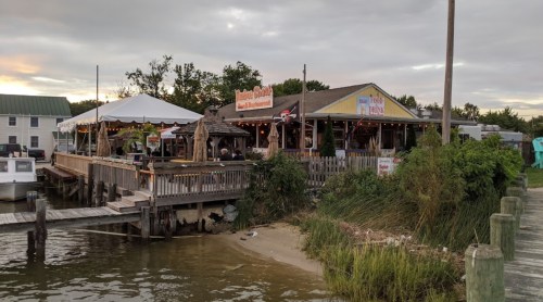 A waterfront restaurant with outdoor seating, surrounded by greenery and a cloudy sky. Boats are docked nearby.