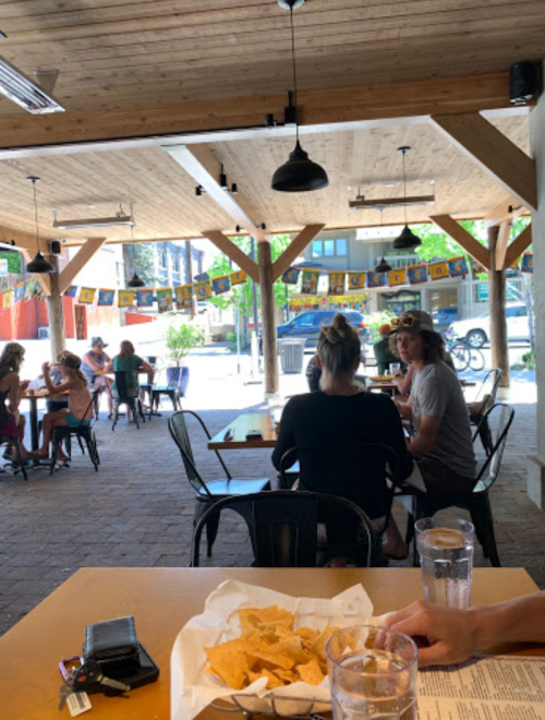 A casual dining space with people sitting at tables, enjoying food and drinks under a wooden roof with colorful decorations.