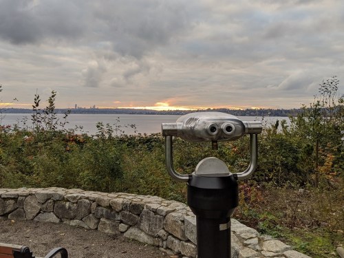 A telescope overlooks a scenic view of a lake and city skyline under a cloudy sky at sunset.
