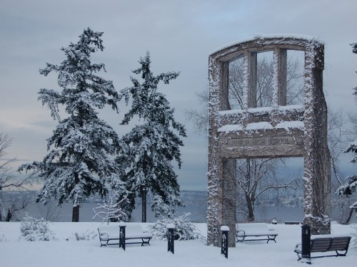 A snowy landscape featuring a stone monument and tall evergreen trees, with a lake visible in the background.