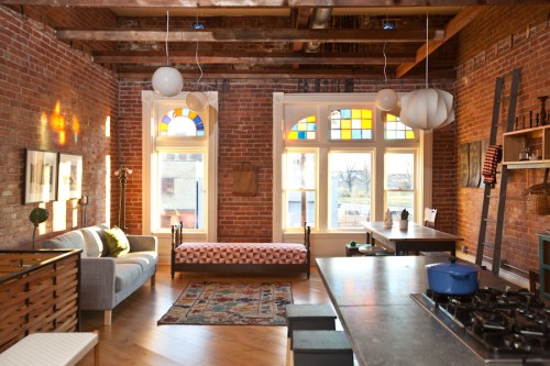 Cozy living space with exposed brick walls, colorful windows, a sofa, and a kitchen area featuring a blue stove.