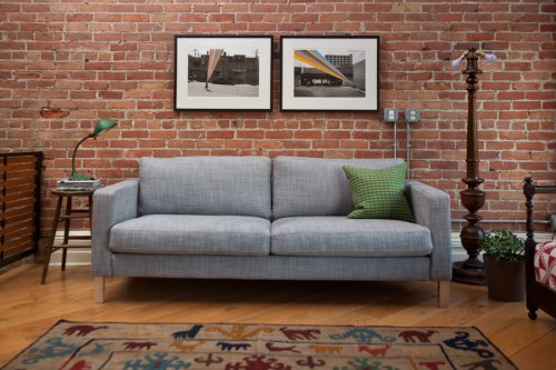 A modern gray sofa with a green pillow, framed photos on a brick wall, and a vintage lamp beside a colorful rug.