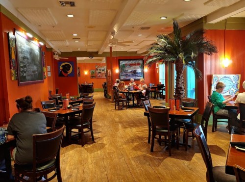 A vibrant restaurant interior with wooden floors, colorful walls, and patrons dining at tables. Palm trees add a tropical touch.