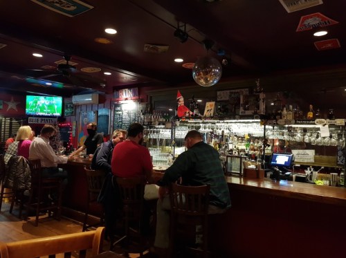 A cozy bar scene with patrons seated at the counter, a TV showing a game, and festive decorations on the walls.