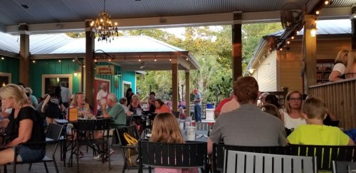 A lively outdoor dining area with people seated at tables, surrounded by greenery and warm lighting.
