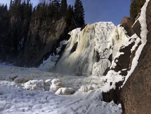 A frozen waterfall cascading over rocky cliffs, surrounded by snow and ice, under a clear blue sky.