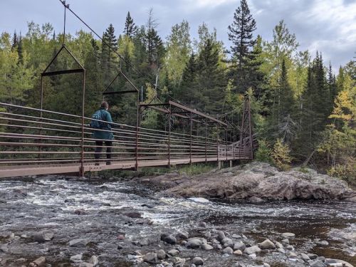 A person stands on a metal suspension bridge over a river, surrounded by lush green trees and rocky terrain.