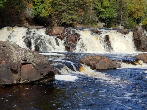 A serene waterfall cascades over rocky ledges, surrounded by lush greenery and autumn-colored trees.