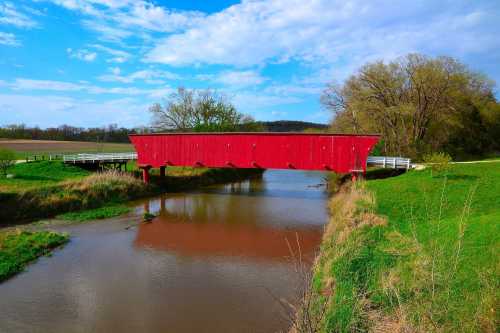 A red covered bridge spans a calm river, surrounded by green fields and trees under a blue sky with clouds.