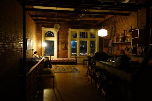 A dimly lit room with exposed brick walls, a cozy seating area, and large windows showing a nighttime view.