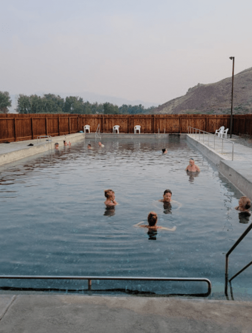 A group of people swimming in a large pool surrounded by a wooden fence and hills in the background.