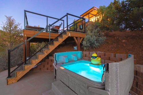 A hot tub with a rubber duck floats inside, surrounded by a wooden deck and steps leading to a house in the background.