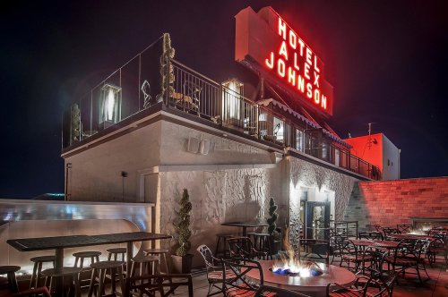 A rooftop view of the Hotel Alex Johnson at night, featuring a glowing sign and outdoor seating around a fire pit.