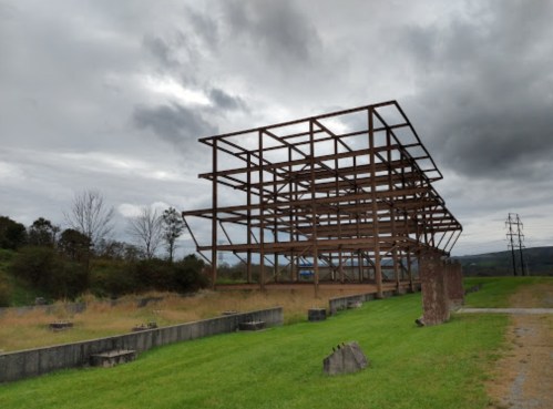 A skeletal metal structure stands in a grassy area under a cloudy sky, with remnants of a foundation nearby.