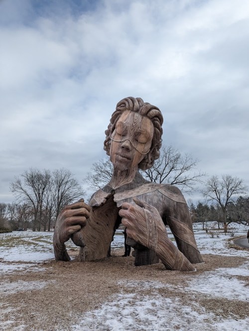 A large wooden sculpture of a person with closed eyes, emerging from the ground, surrounded by a snowy landscape.