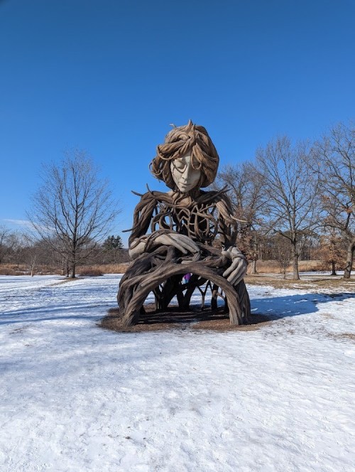 A large, artistic sculpture of a figure with tree-like features, surrounded by snow and bare trees under a clear blue sky.