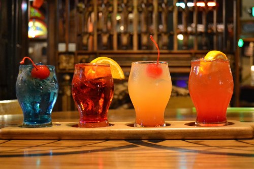 Four colorful cocktails on a wooden tray, featuring cherries and orange slices, set against a bar backdrop.