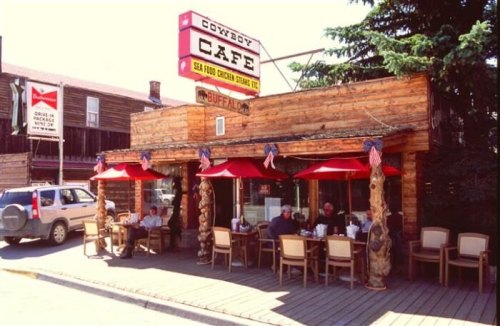 A rustic café with outdoor seating, featuring red umbrellas and American flags, surrounded by trees and a sunny sky.