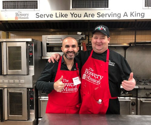 Two men in red aprons with "The Bowery Mission" logo smile and give thumbs up in a kitchen setting.