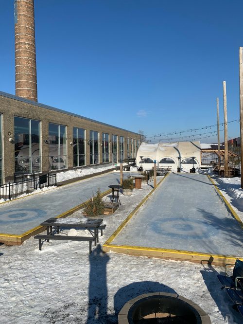 Outdoor ice skating rink with clear blue skies, surrounded by tables and a building, featuring a tall chimney in the background.